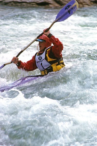 Kayaker in red jacket navigates white water rapids, holding paddle, wearing life vest, number bib.