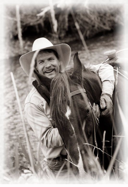 Cowboy hugging brown horse, smiling, wearing hat and gloves outdoors.