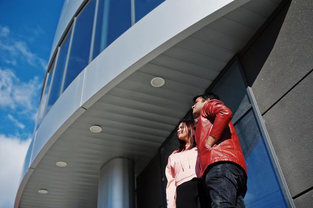 Two people looking up at a modern building, one in a red jacket, the other in a pink sweater, against a blue sky.