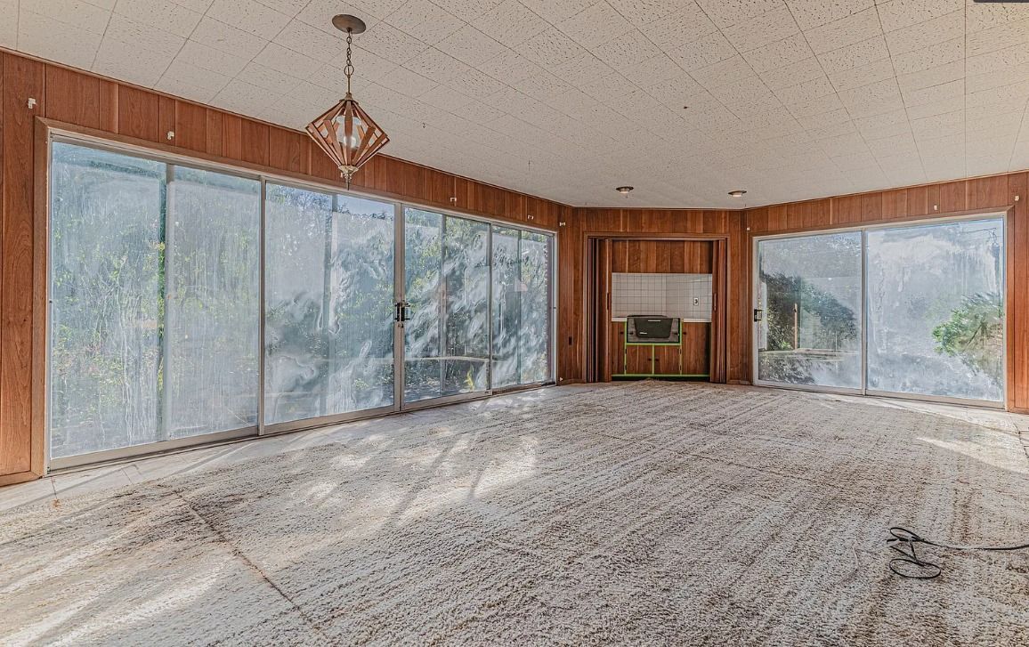 Empty room with dirty carpet, wood paneling, large windows, and a chandelier.