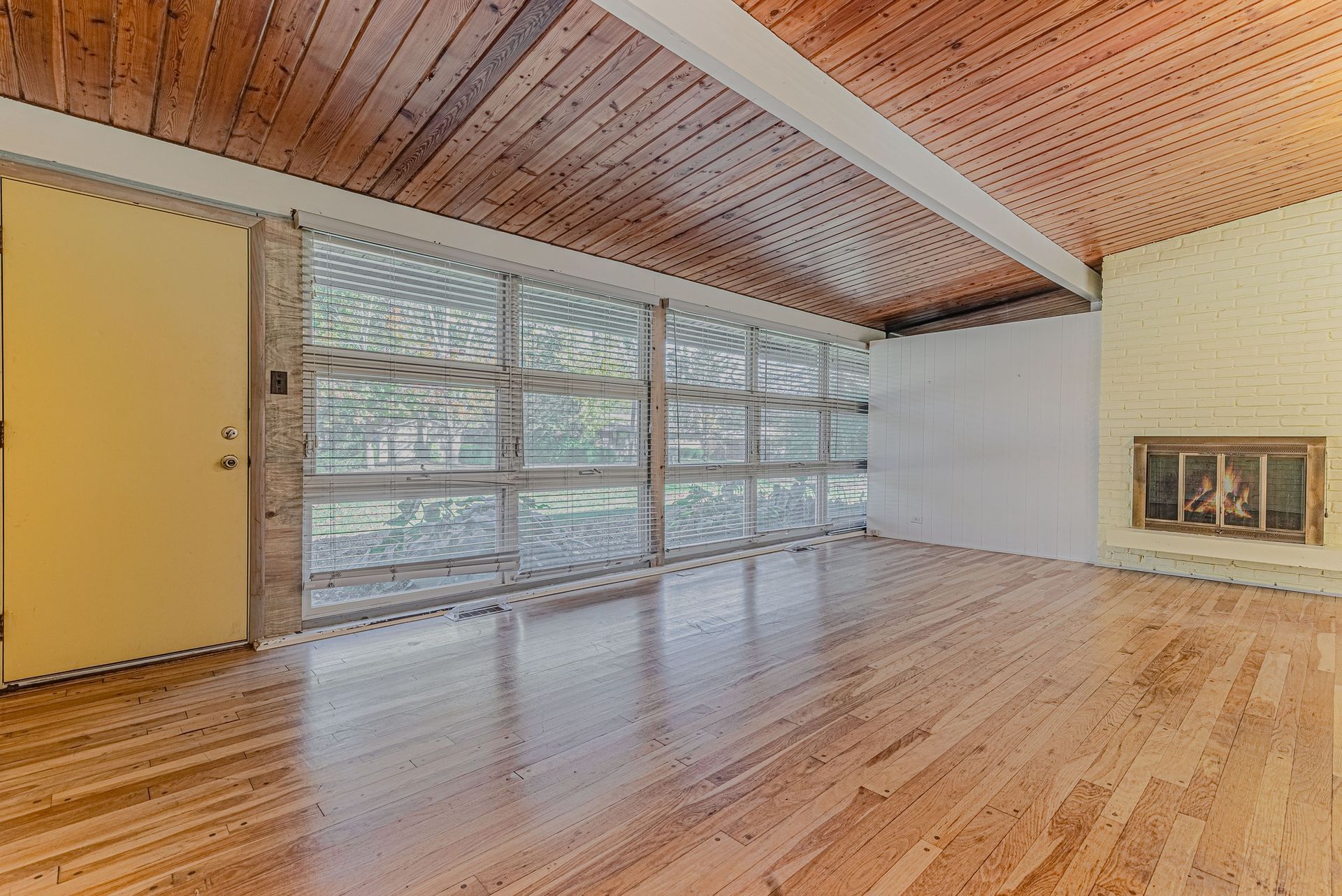 Empty room with wood floors, ceiling, and large windows. Fireplace on right. Yellow door on left.