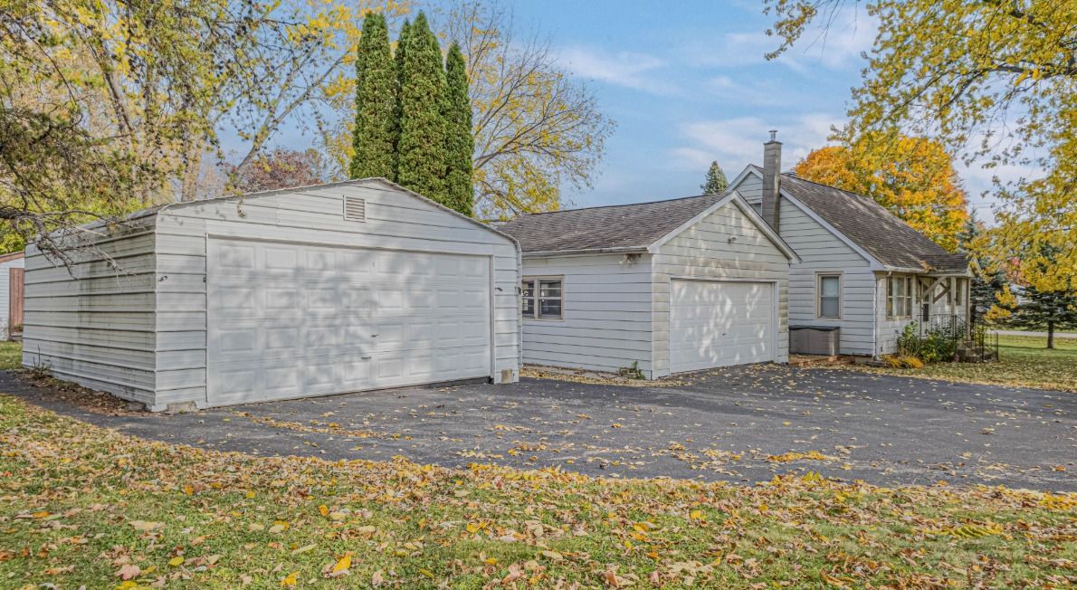 White house with two-car garage on a leaf-covered driveway, surrounded by trees.