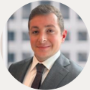 Headshot of a person wearing a grey suit and dark tie, smiling softly against a blurred office building background.