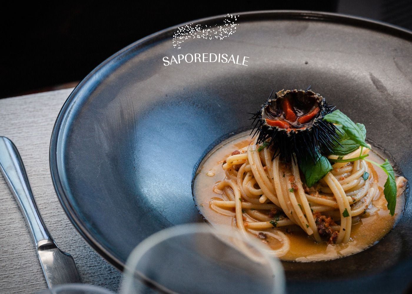 Piatto di pasta con contorno di ricci di mare in una ciotola scura, accanto a una forchetta e un bicchiere.