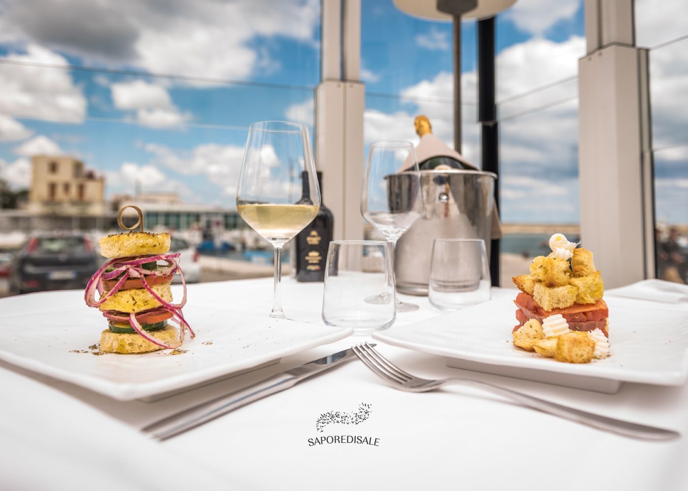 Due antipasti impiattati, bicchieri di vino e acqua su un tavolo con vista sul porto, cielo azzurro e nuvole.
