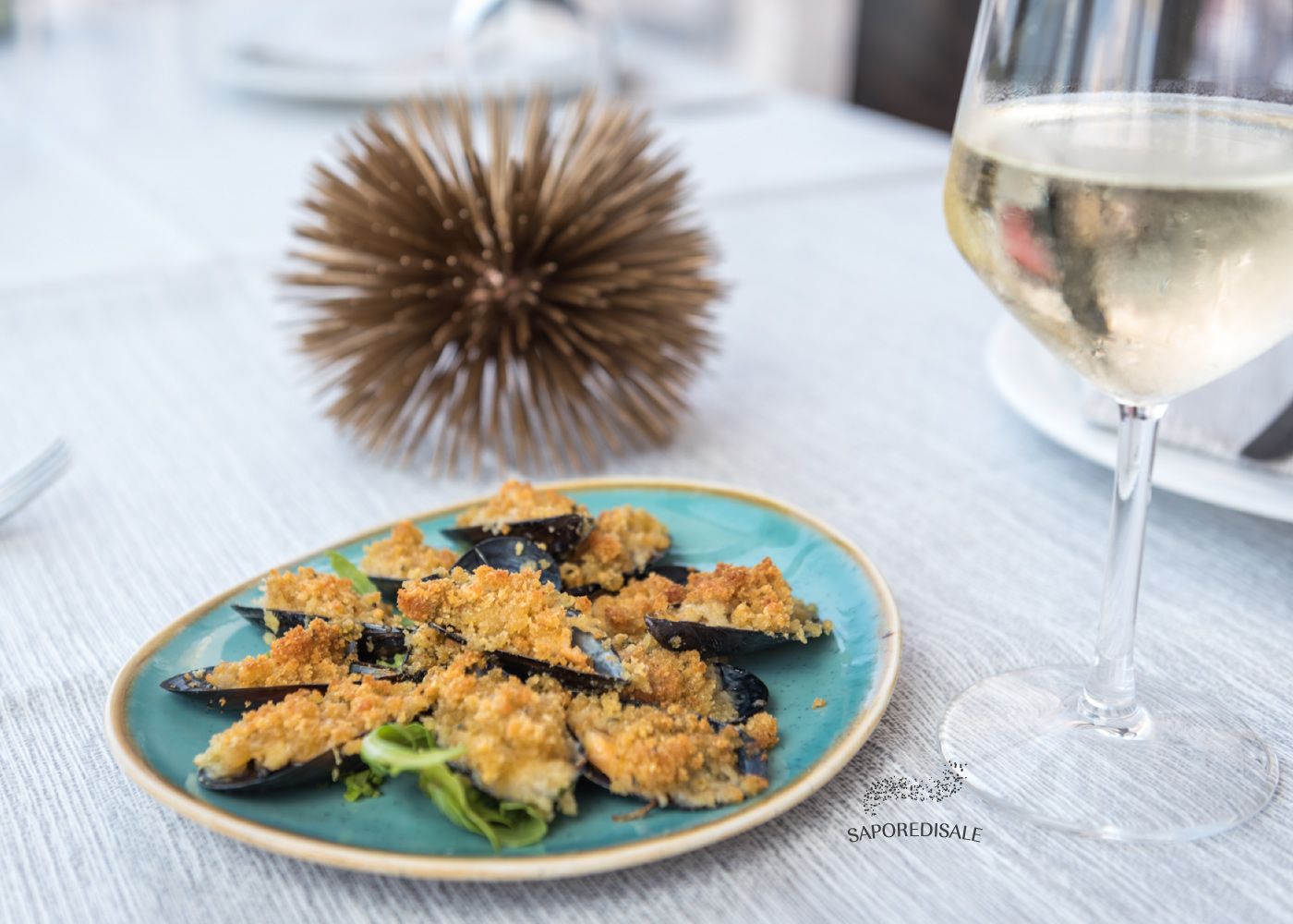 Cozze al forno su un piatto blu, con un bicchiere di vino bianco sul tavolo.