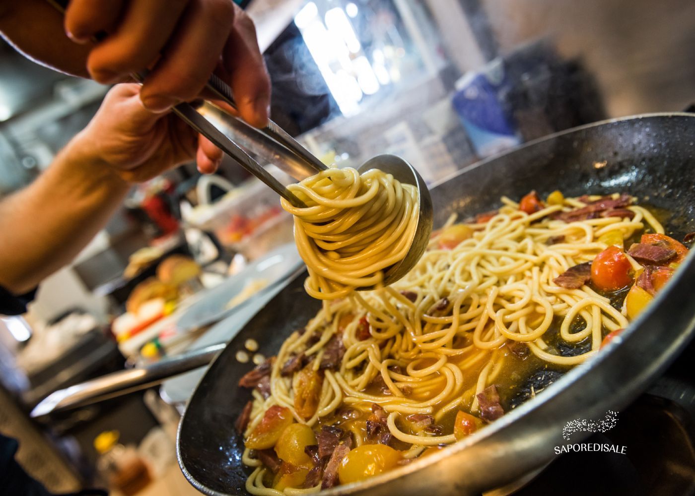 Persona che usa le pinze per condire la pasta con il sugo e i pomodori in una padella sul fornello.