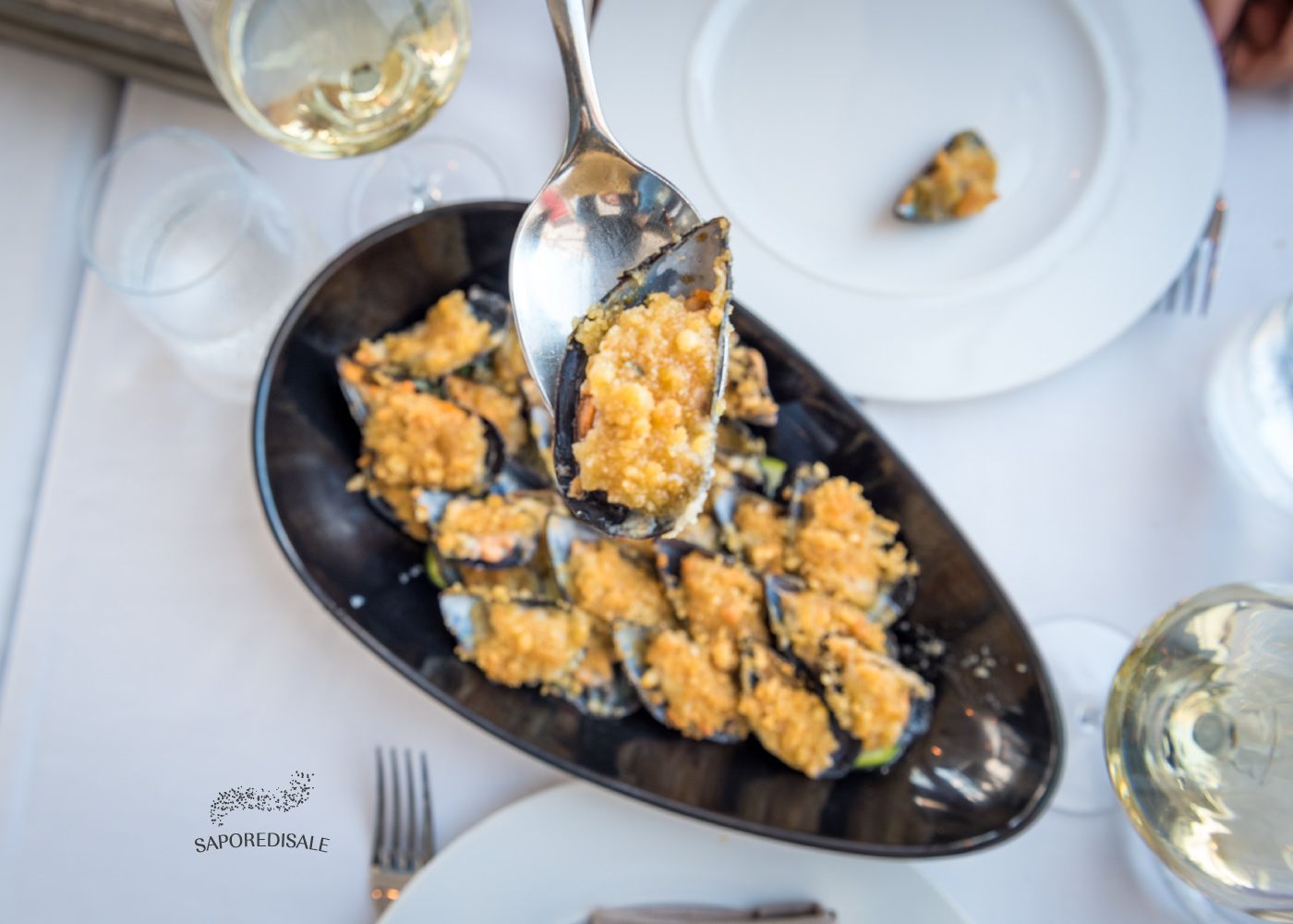 Cozze al forno su un piatto ovale nero. Un cucchiaio ne raccoglie una. Bicchieri e piatti da vino bianco circondano.