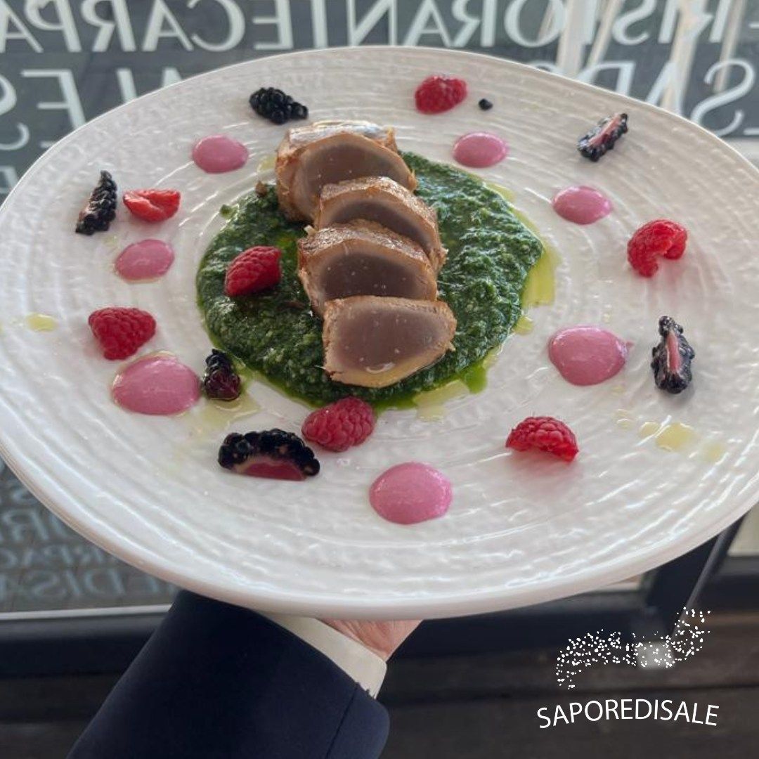 Carne affettata su salsa verde, guarnita con frutti di bosco e pois rosa su un piatto bianco.