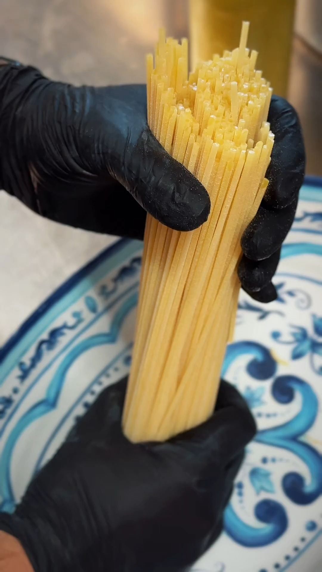 Mani in guanti neri che tengono un fascio di spaghetti crudi su un piatto decorativo blu e bianco.