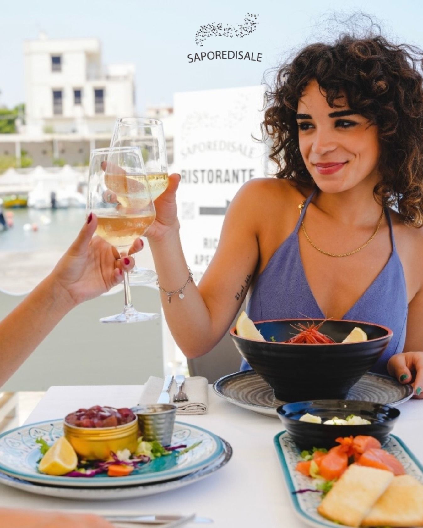 Donna che brinda al vino in un ristorante all'aperto. Piatti di pesce sul tavolo. Il lungomare sullo sfondo.