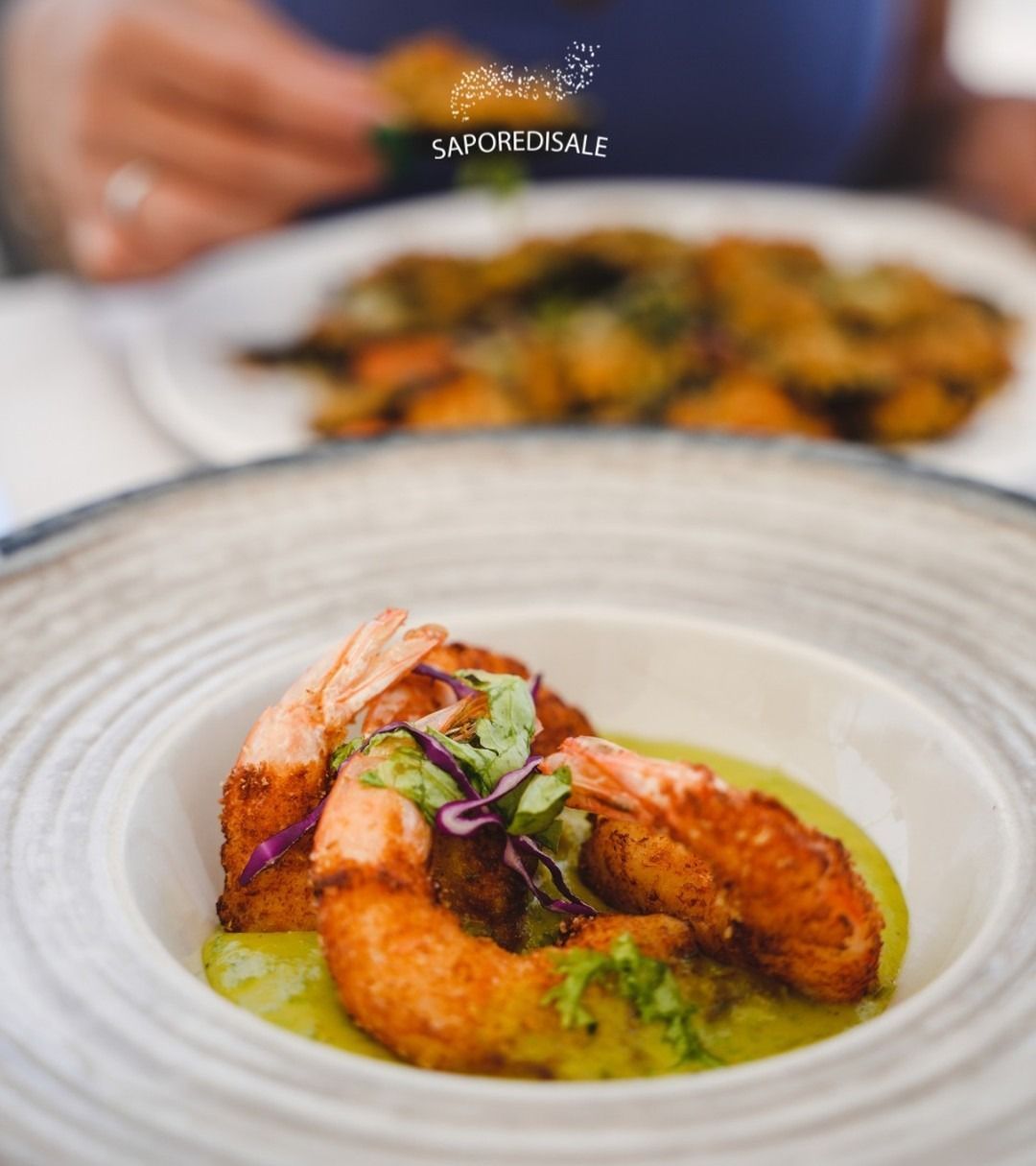 Piatto di gamberi fritti con salsa verde, un altro piatto sullo sfondo. Qualcuno sta mangiando.