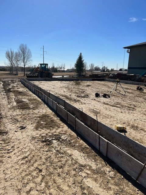 Construction site: Wooden forms set for a concrete foundation on a dirt lot, tractor in background, blue sky.