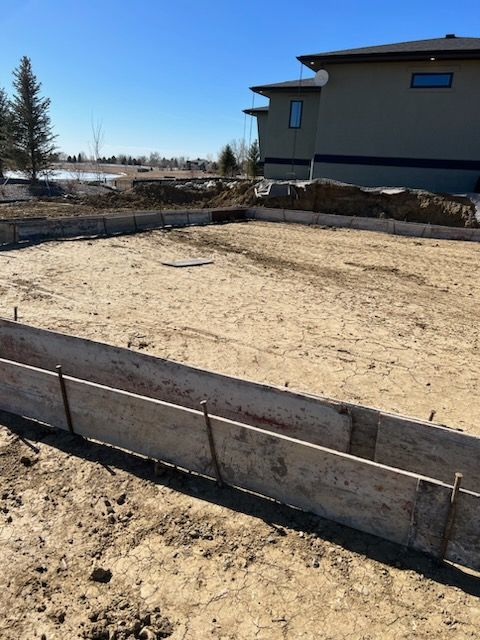 Foundation construction site with wooden forms and a beige house in the background under a blue sky.