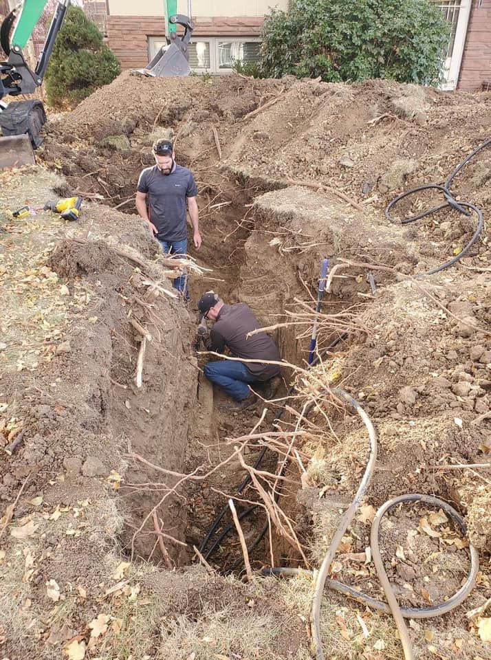 Two men in a deep trench, working on underground pipes, dirt pile in background.