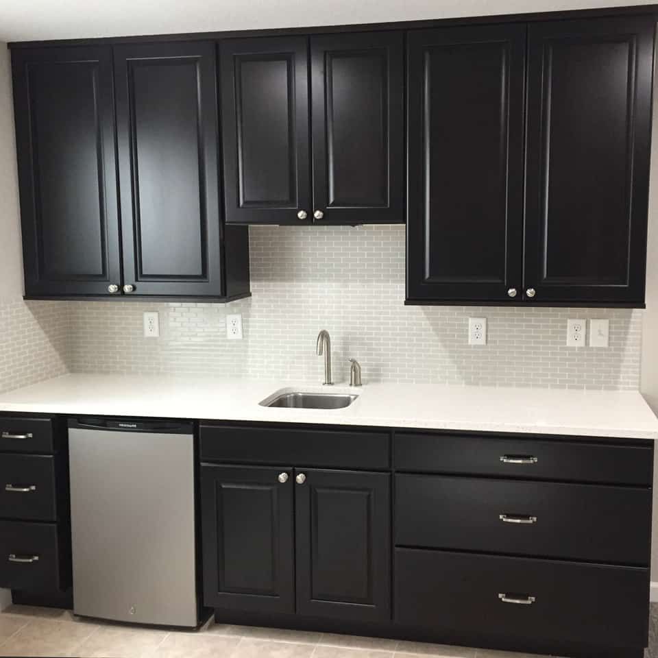 Black cabinets in a kitchen with a white countertop, stainless steel mini fridge, and silver faucet.