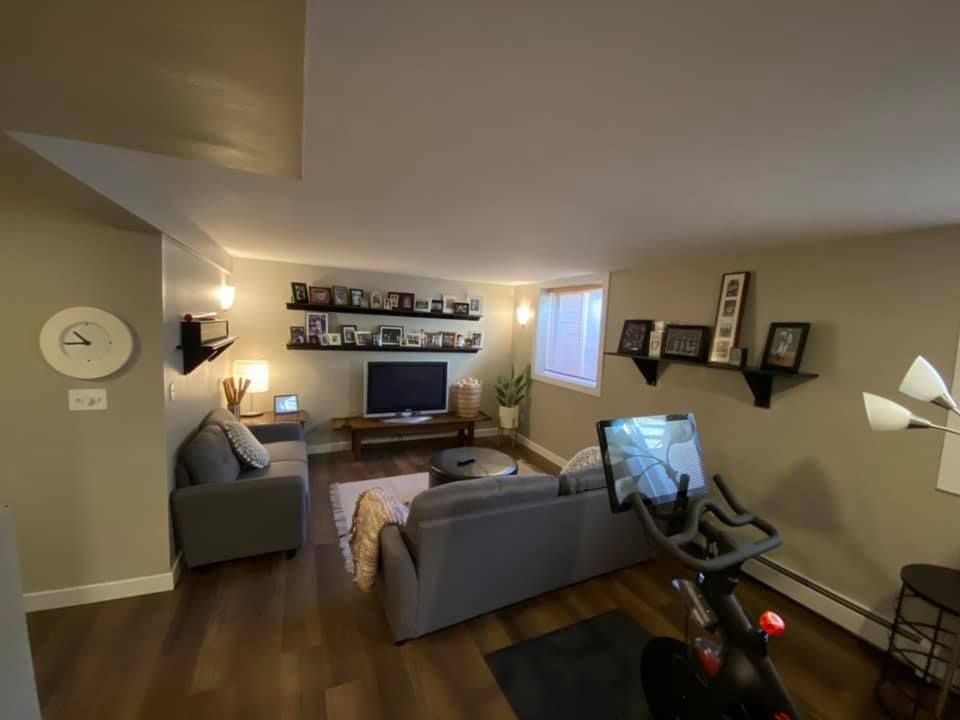 Basement living room with a gray couch, TV, Peloton bike, and photo display.