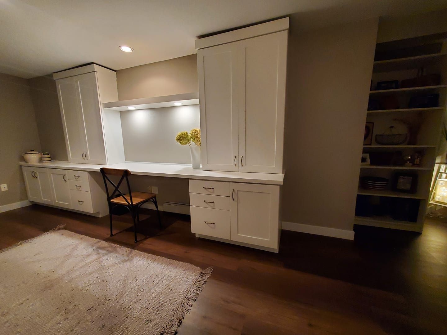 Built-in white desk with cabinets, shelves, and a chair in a room with wood flooring and a rug.