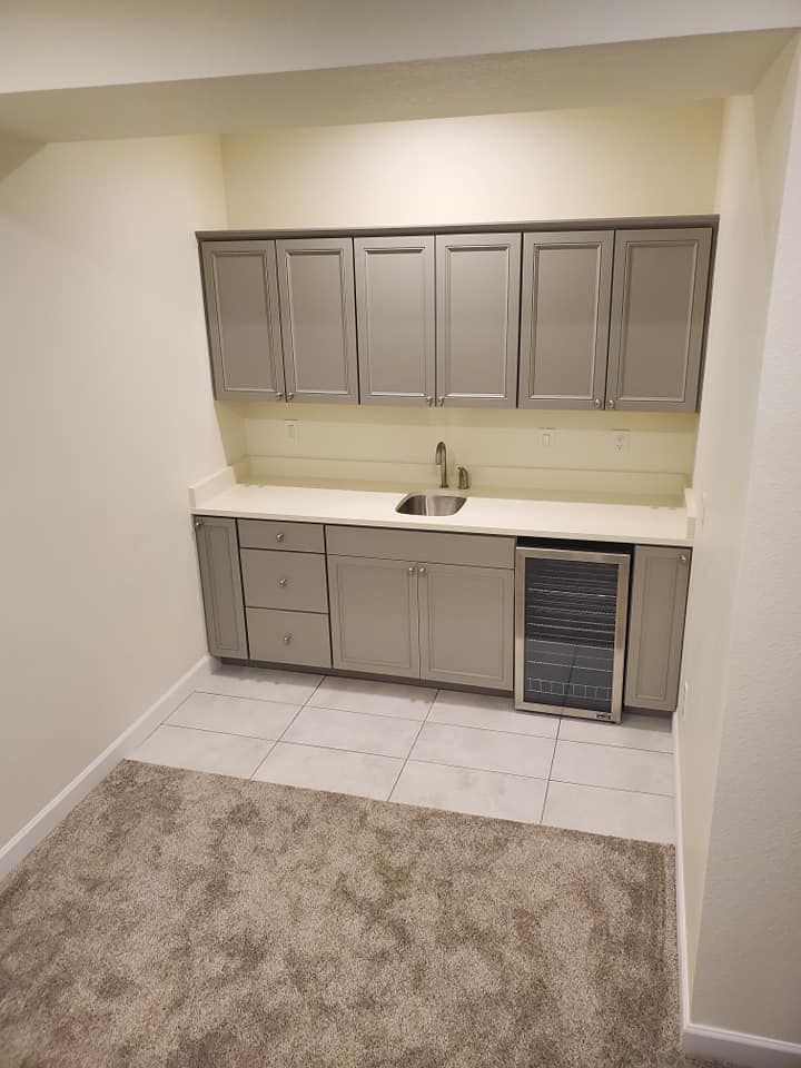 Gray cabinets and countertop with a sink and beverage cooler in a small room with tile and carpet.