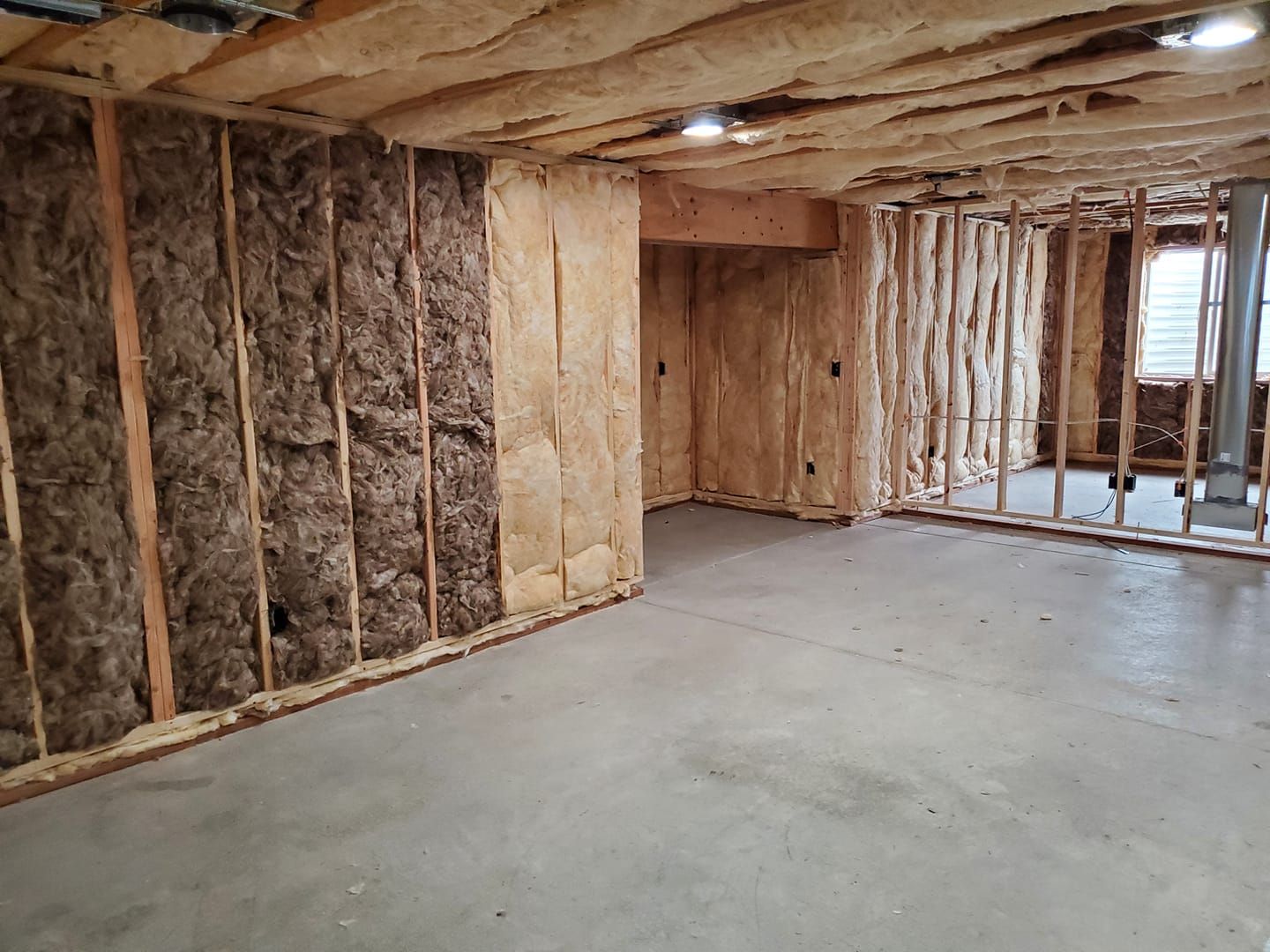 Basement under construction, featuring insulation between wooden studs and on the ceiling, with concrete flooring.