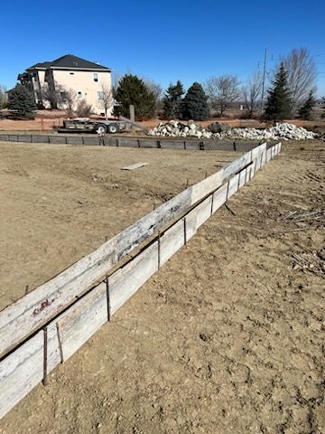 Construction site with wooden forms for a foundation, dirt ground, house in background.