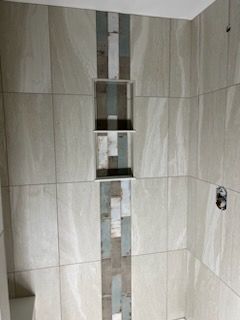 Shower wall with a niche, tiled in neutral colors, with a vertical strip of multi-colored tiles.