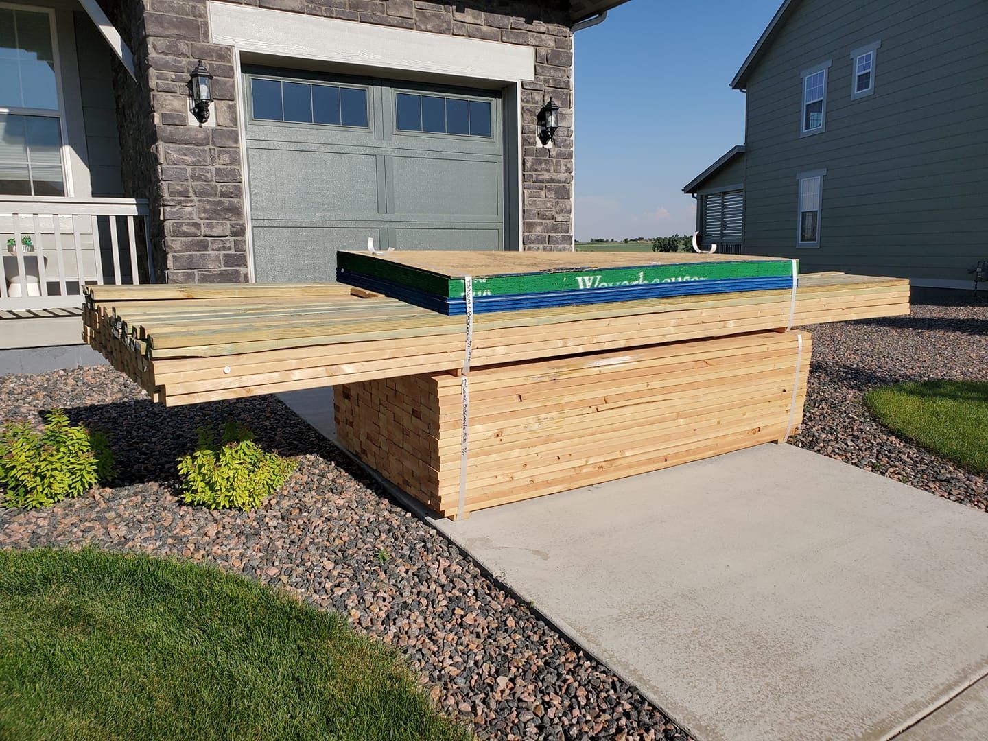 A wooden table with a green and blue box on top sits on a concrete path next to a house and green grass.