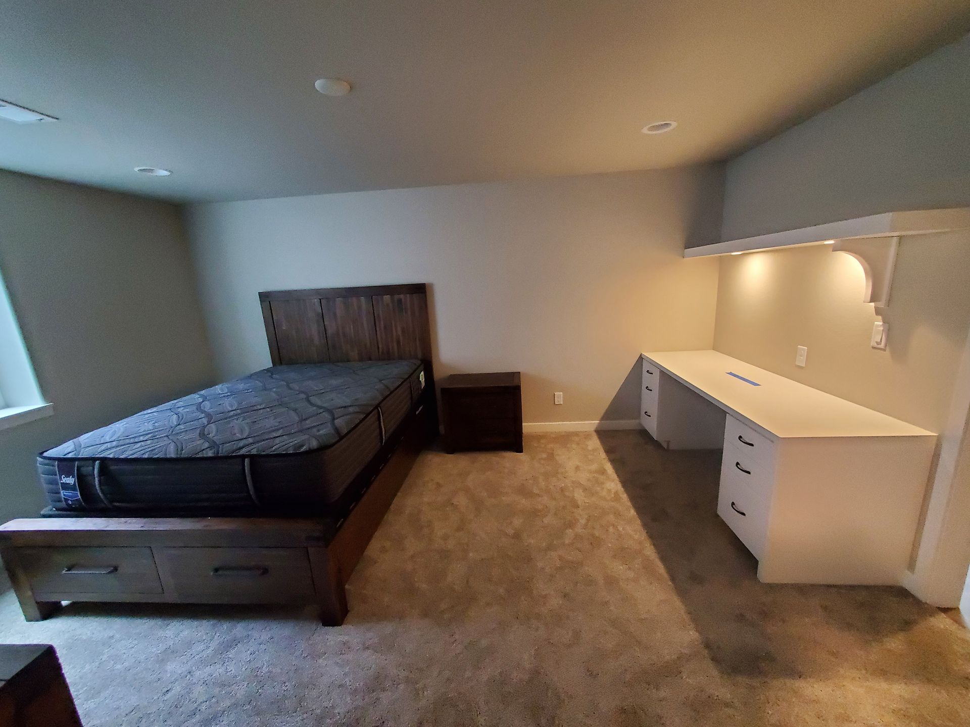 Bedroom with bed, nightstand, and built-in desk with overhead lighting. Beige walls, carpet.