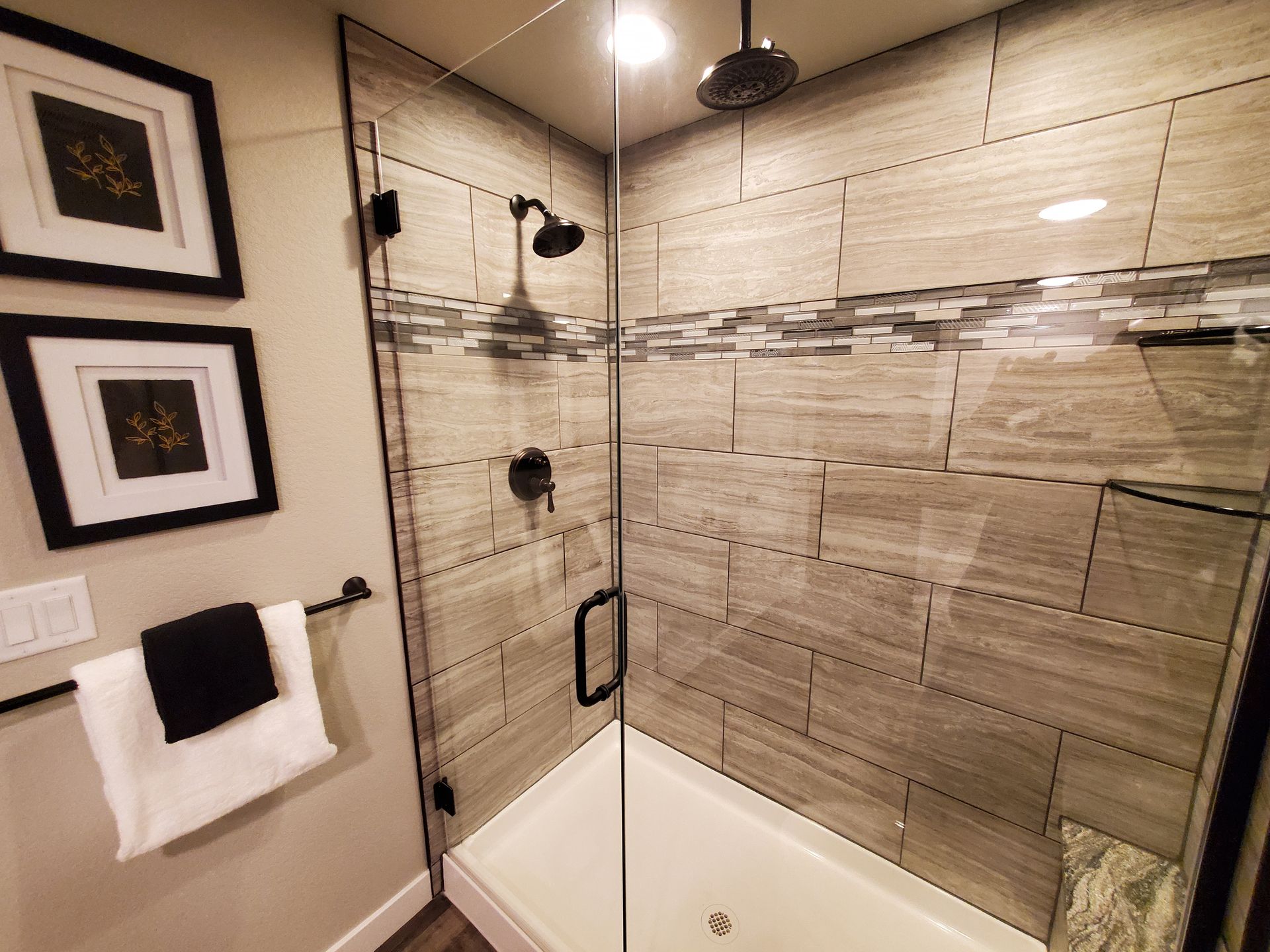 Bathroom with glass shower, gray tile, black fixtures, white tub, artwork, and towel rack.