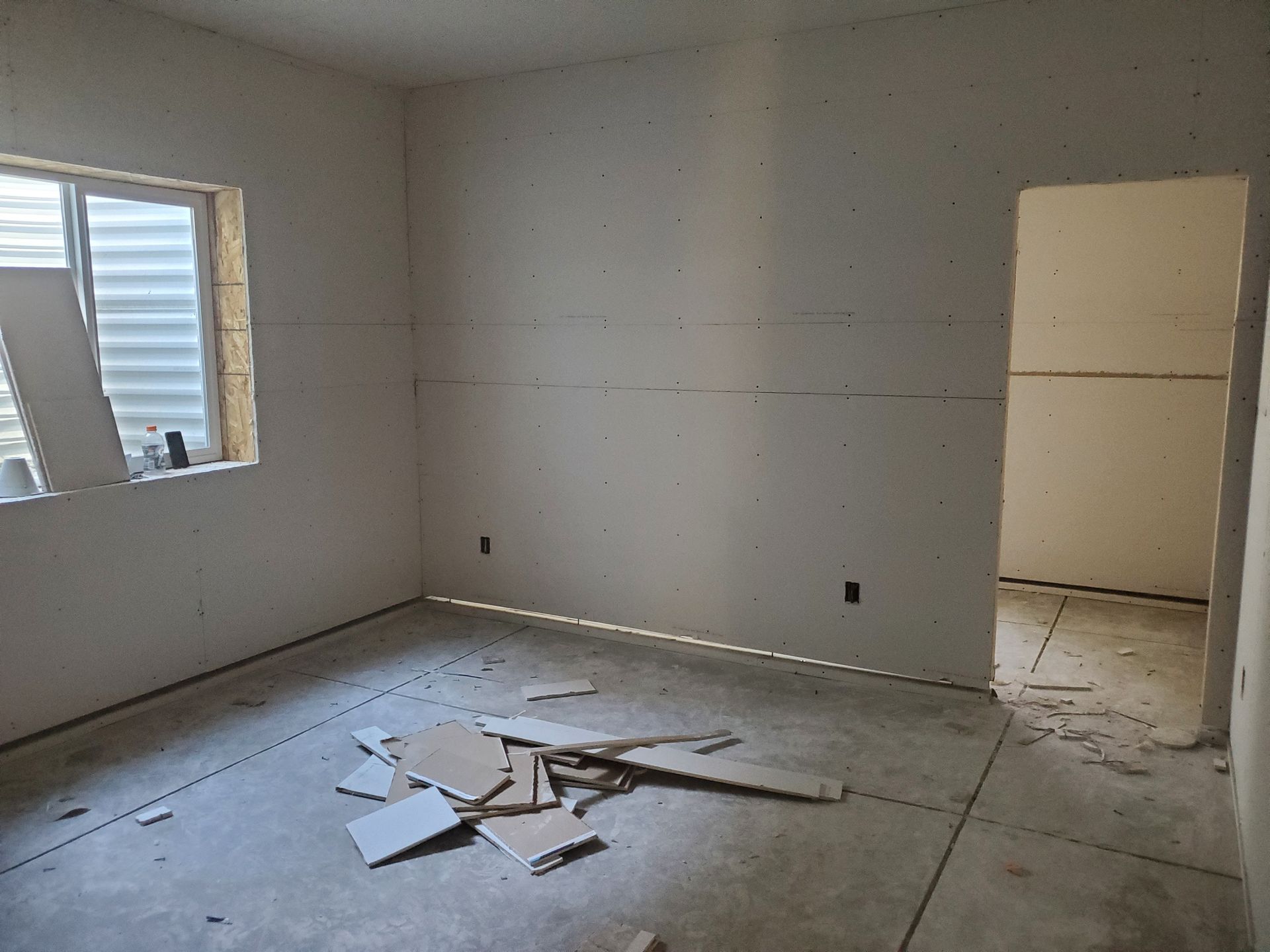 Room with drywall on walls, unfinished floor, debris. Window on the left, doorway on the right.