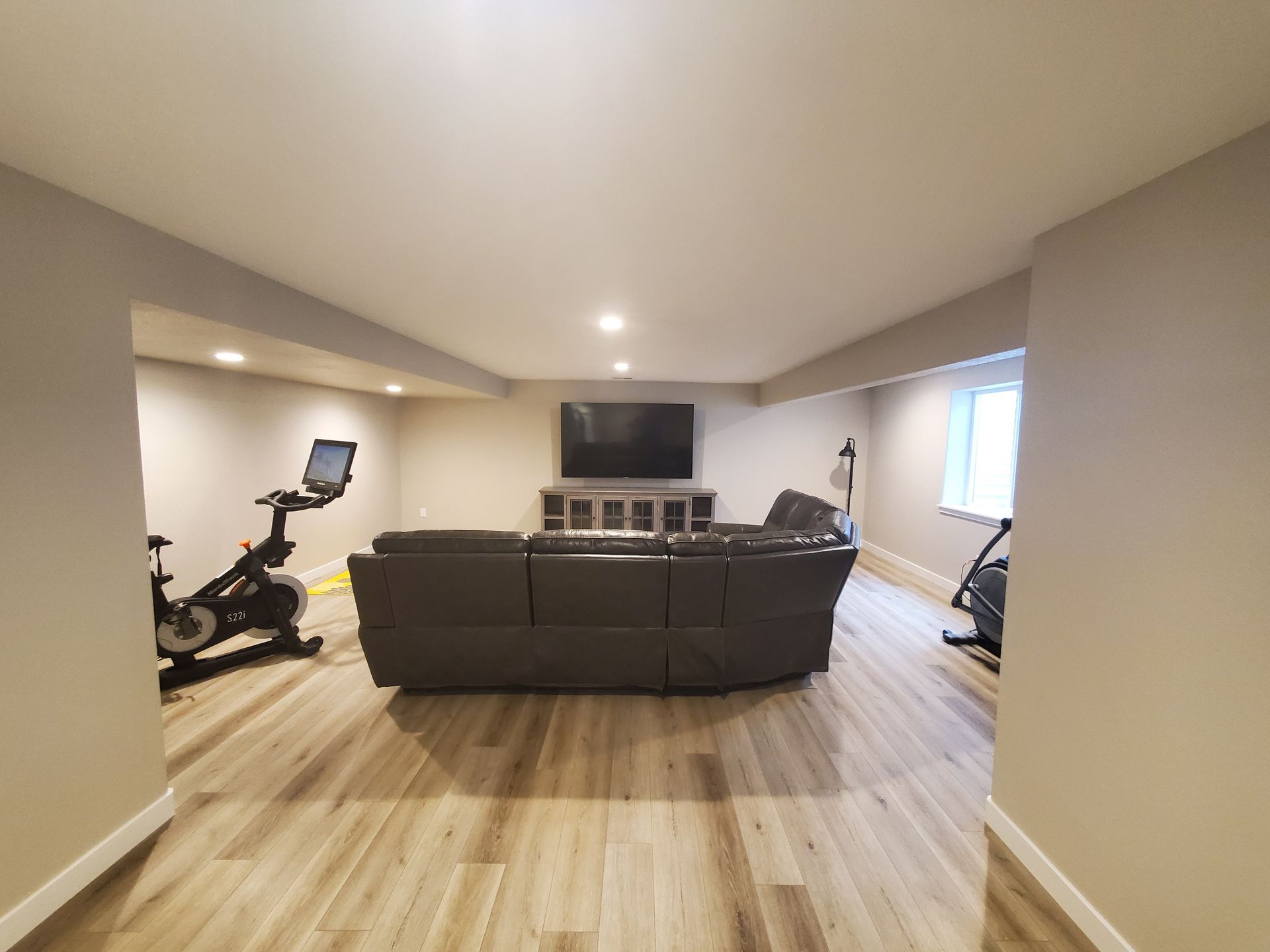 Basement with a Peloton bike, sofa, TV, and exercise equipment. Neutral colors and light wood floors.