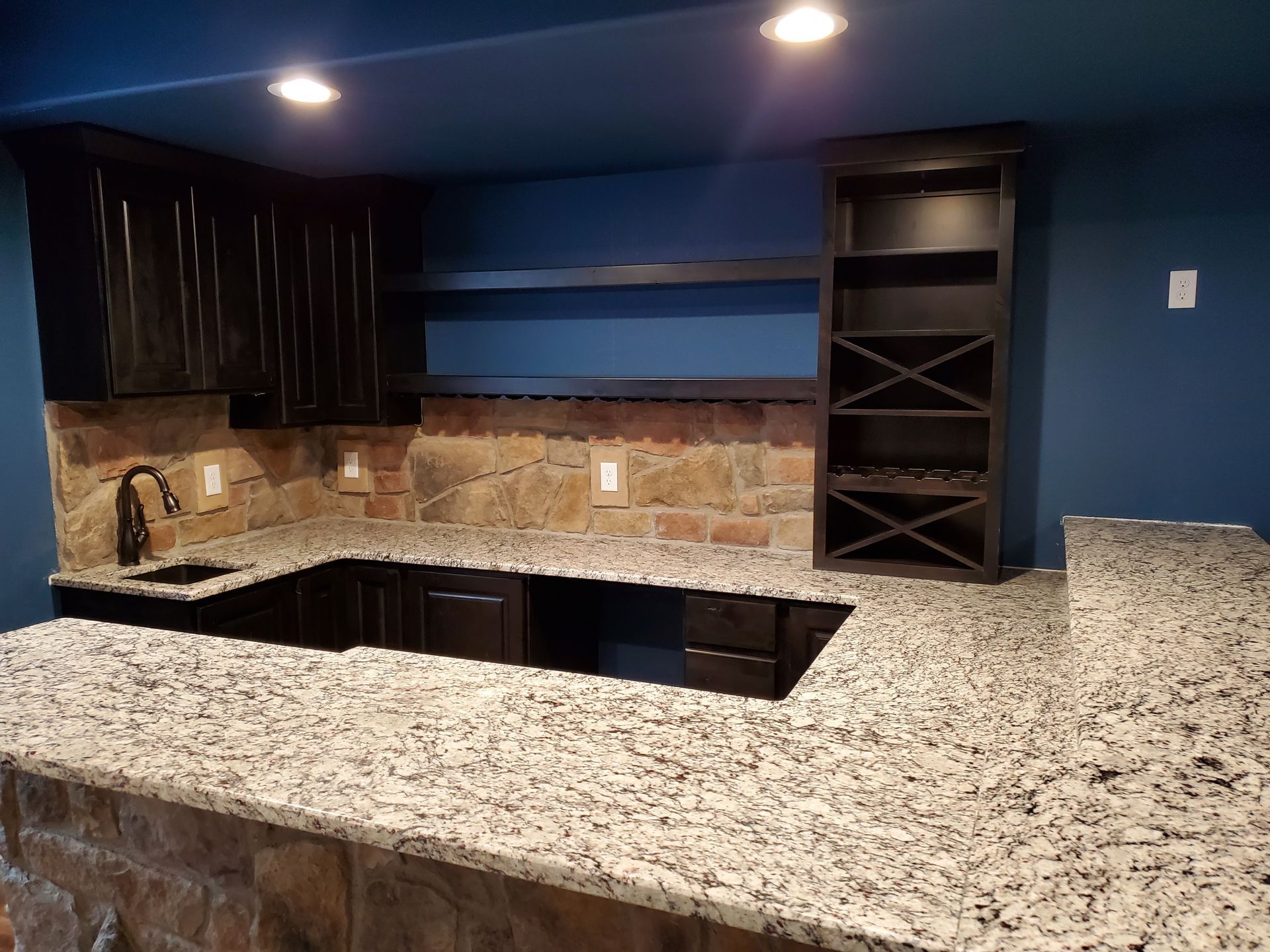 Basement bar with dark cabinets, granite countertop, stone backsplash, and dark blue walls.