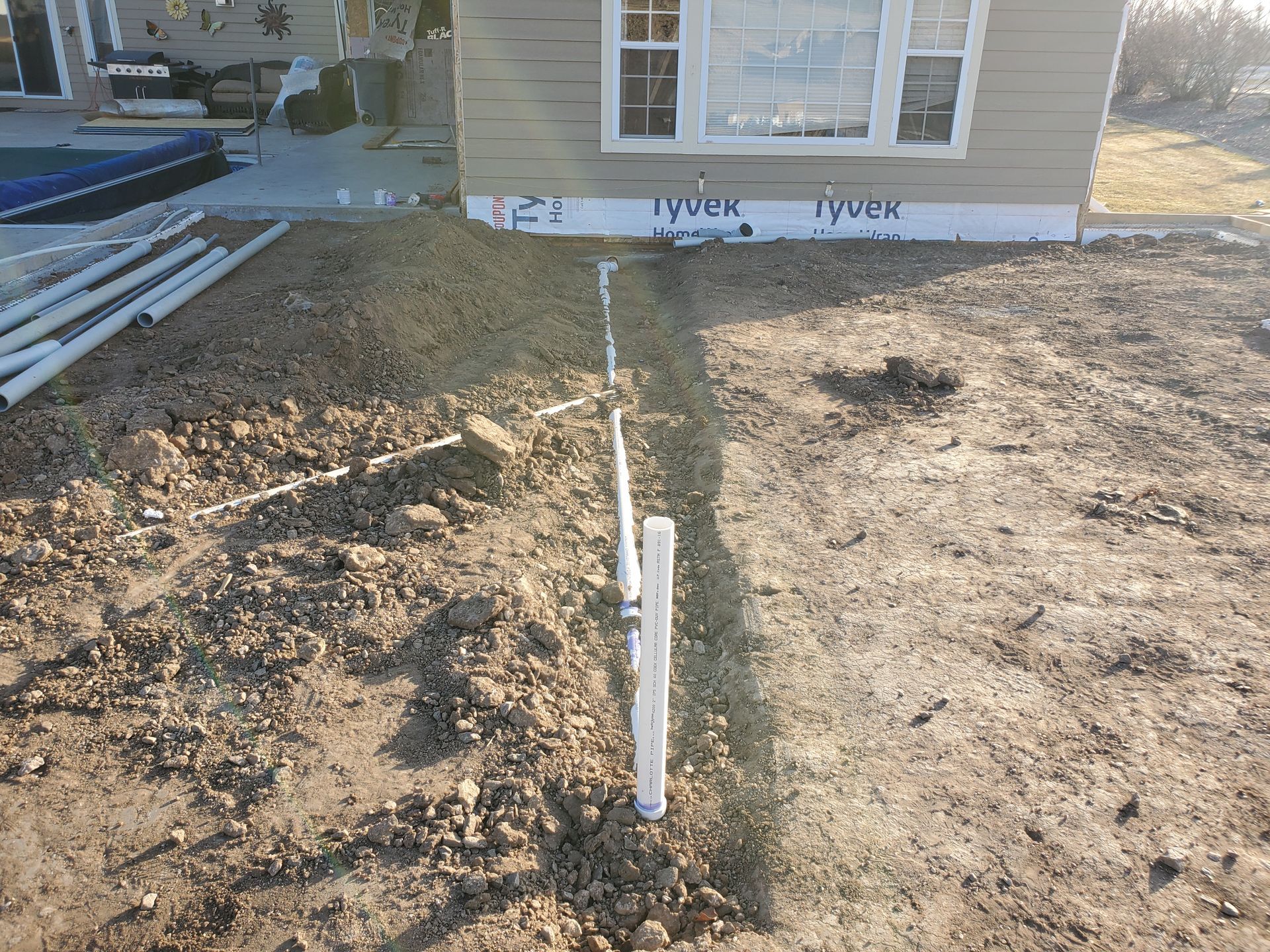 A trench with white PVC pipes laid in the ground in front of a house.