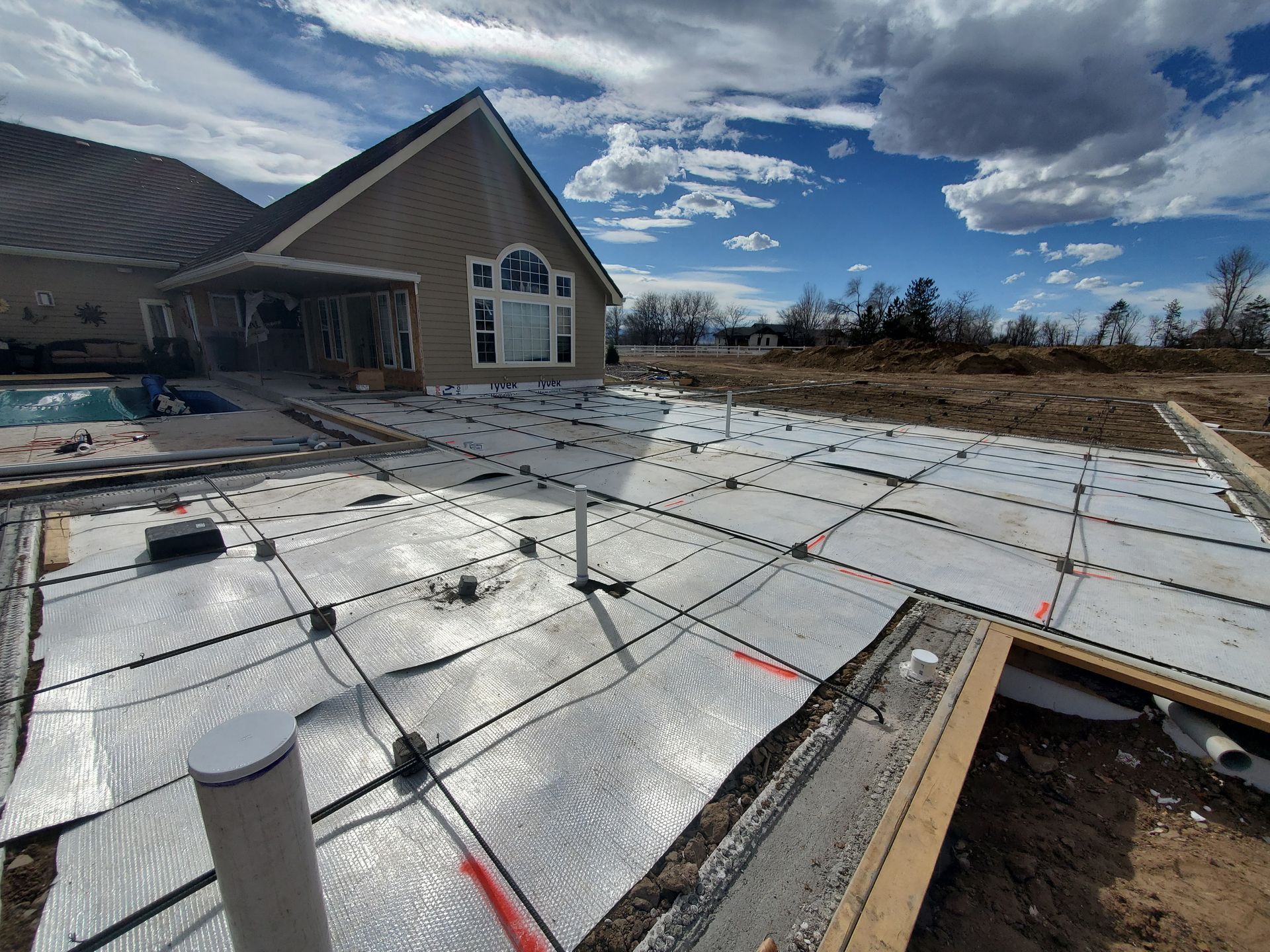 Construction site with concrete foundation and pipes; house in the background; blue sky and clouds.