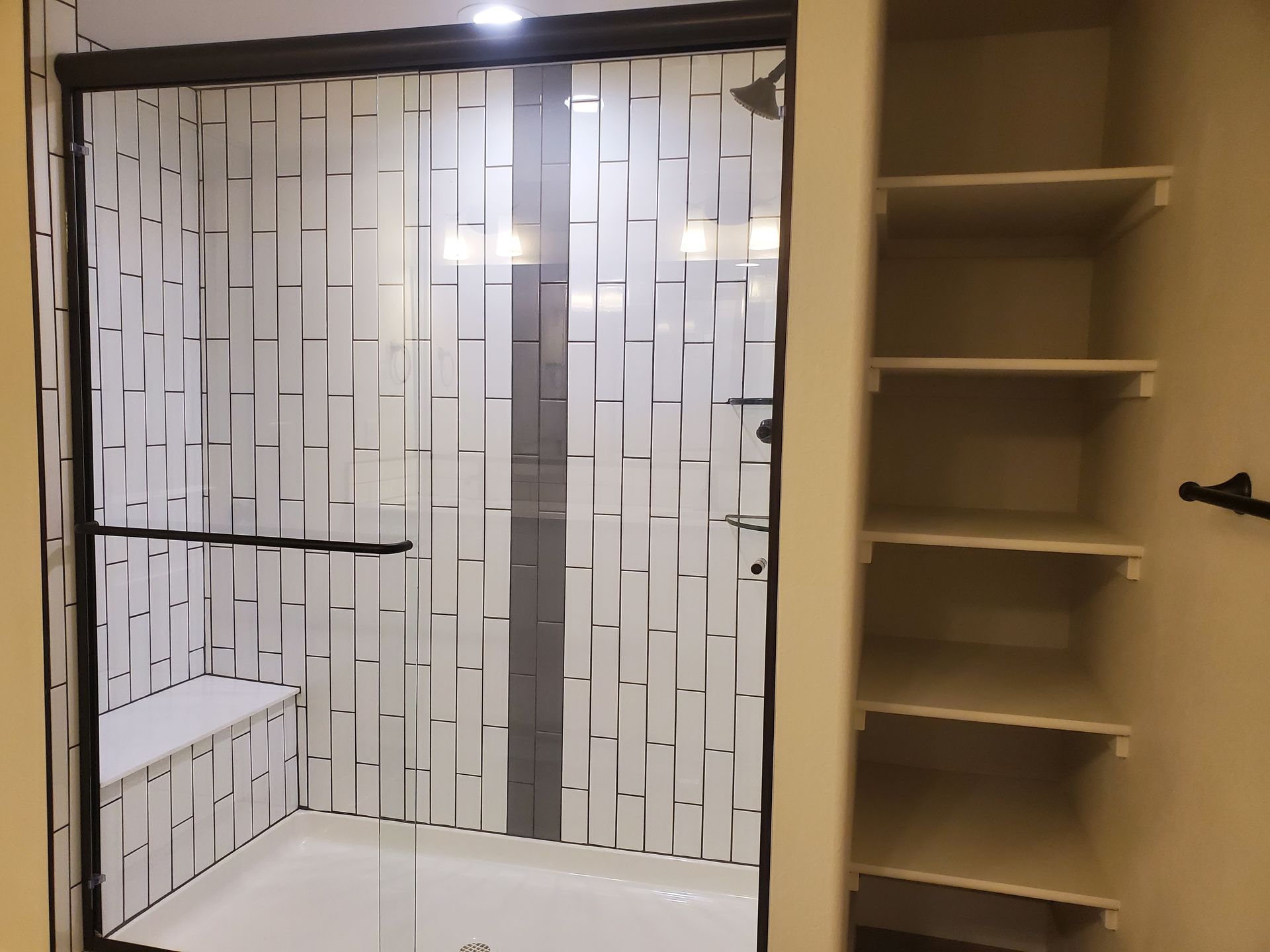 Shower with glass door, tile walls, built-in seat, and adjacent shelving unit in bathroom.