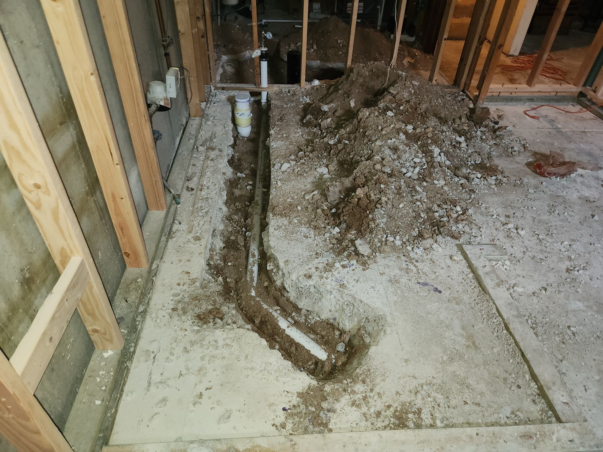 Trench dug in concrete floor with exposed white plumbing pipes, dirt pile nearby.  Framed walls in the background.