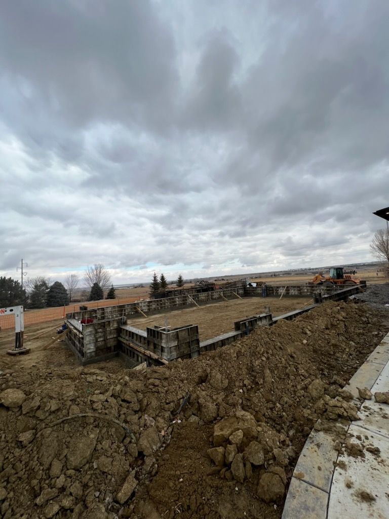 Construction site with foundation walls, dirt, and an excavator under a cloudy sky.