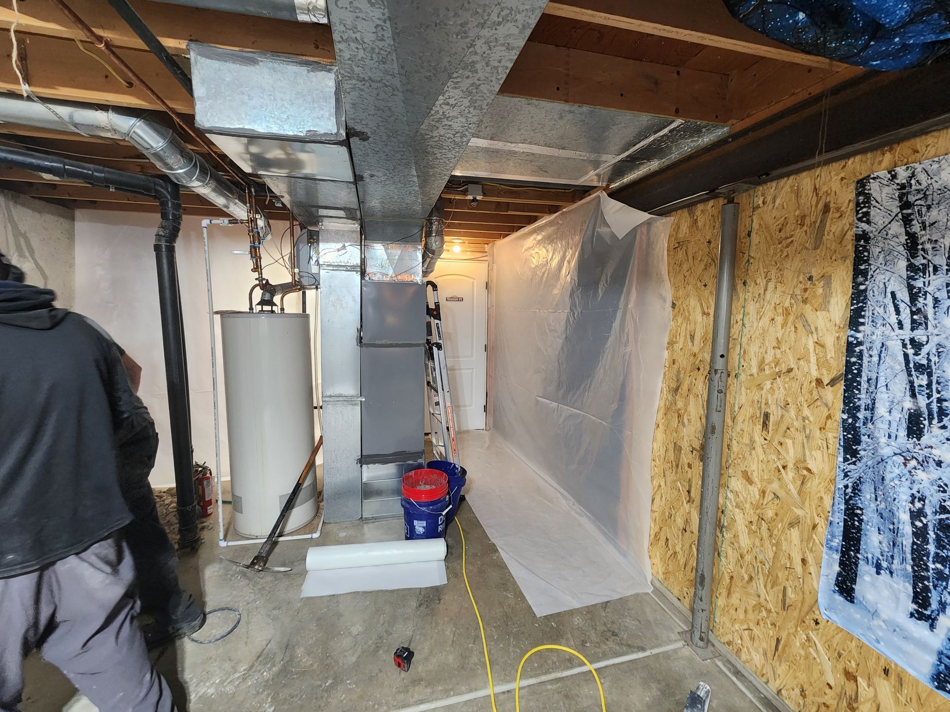 Basement with HVAC, water heater, and plastic sheeting, person on the left. Concrete floor, exposed ceiling.