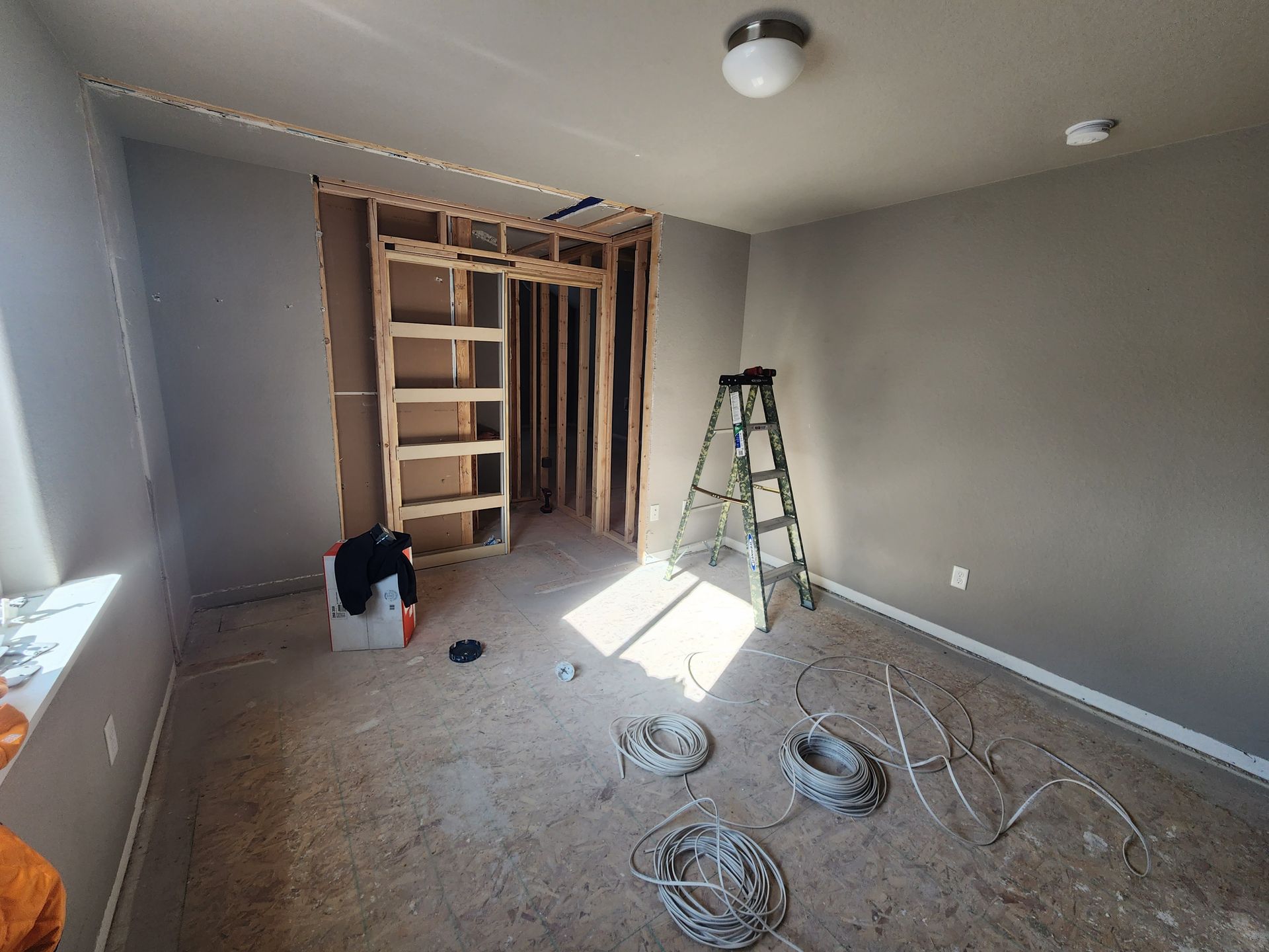 Room under construction; wooden frame, gray walls, ladder, exposed wiring, and a doorway.