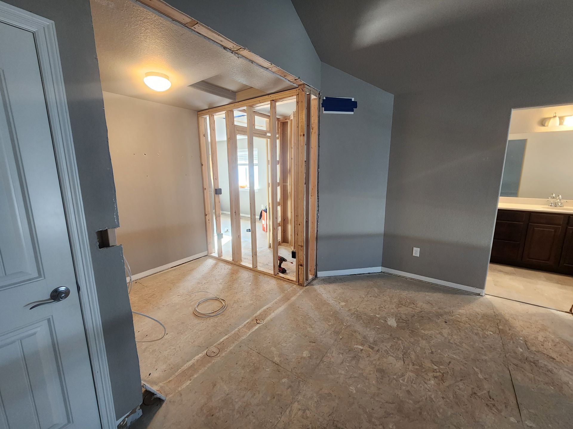 Interior view of a room under construction, featuring exposed wood framing, gray walls, and a view into a bathroom.