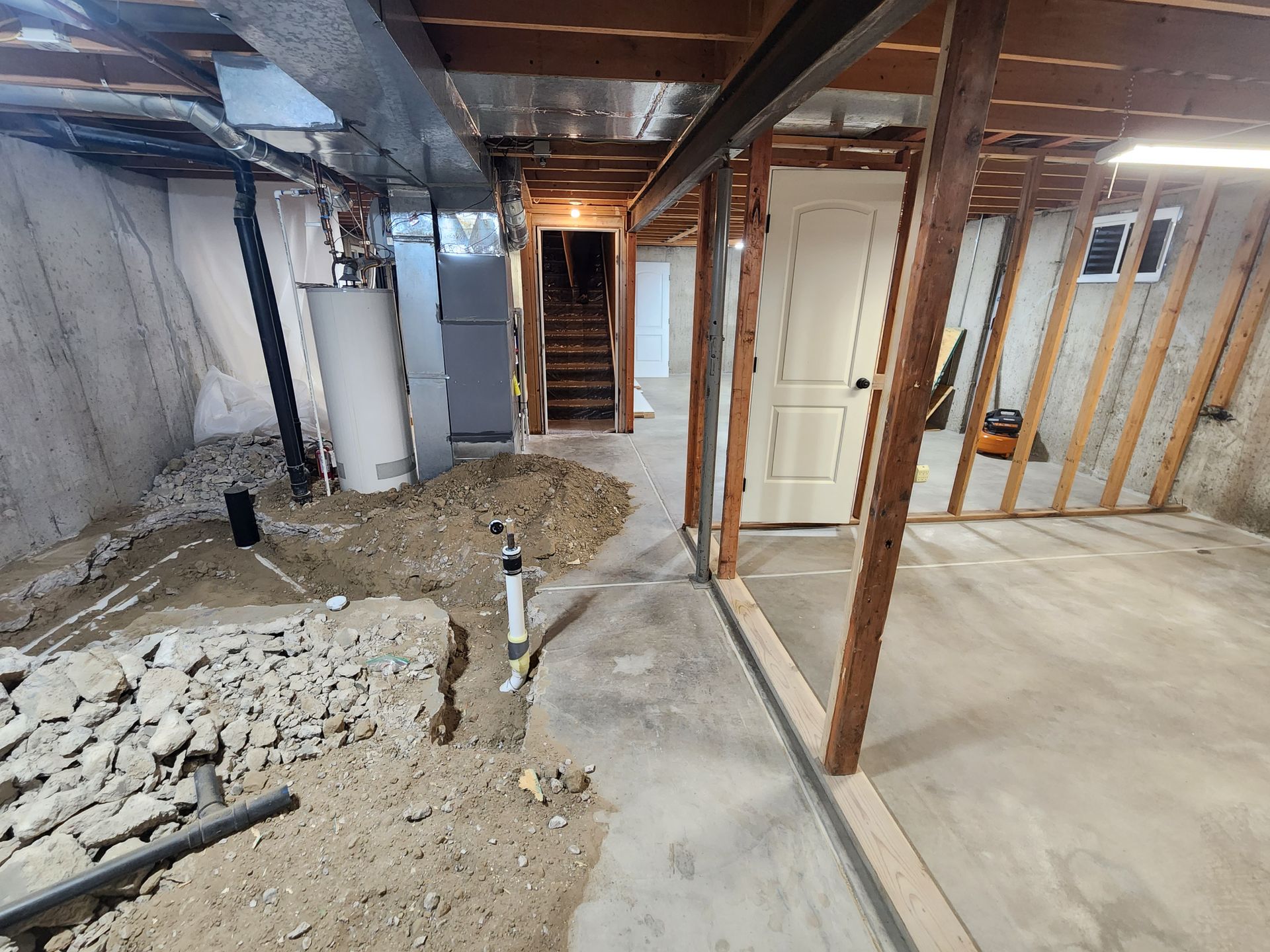 Basement renovation: exposed studs, concrete floor, water heater, rough plumbing, staircase, and a door.