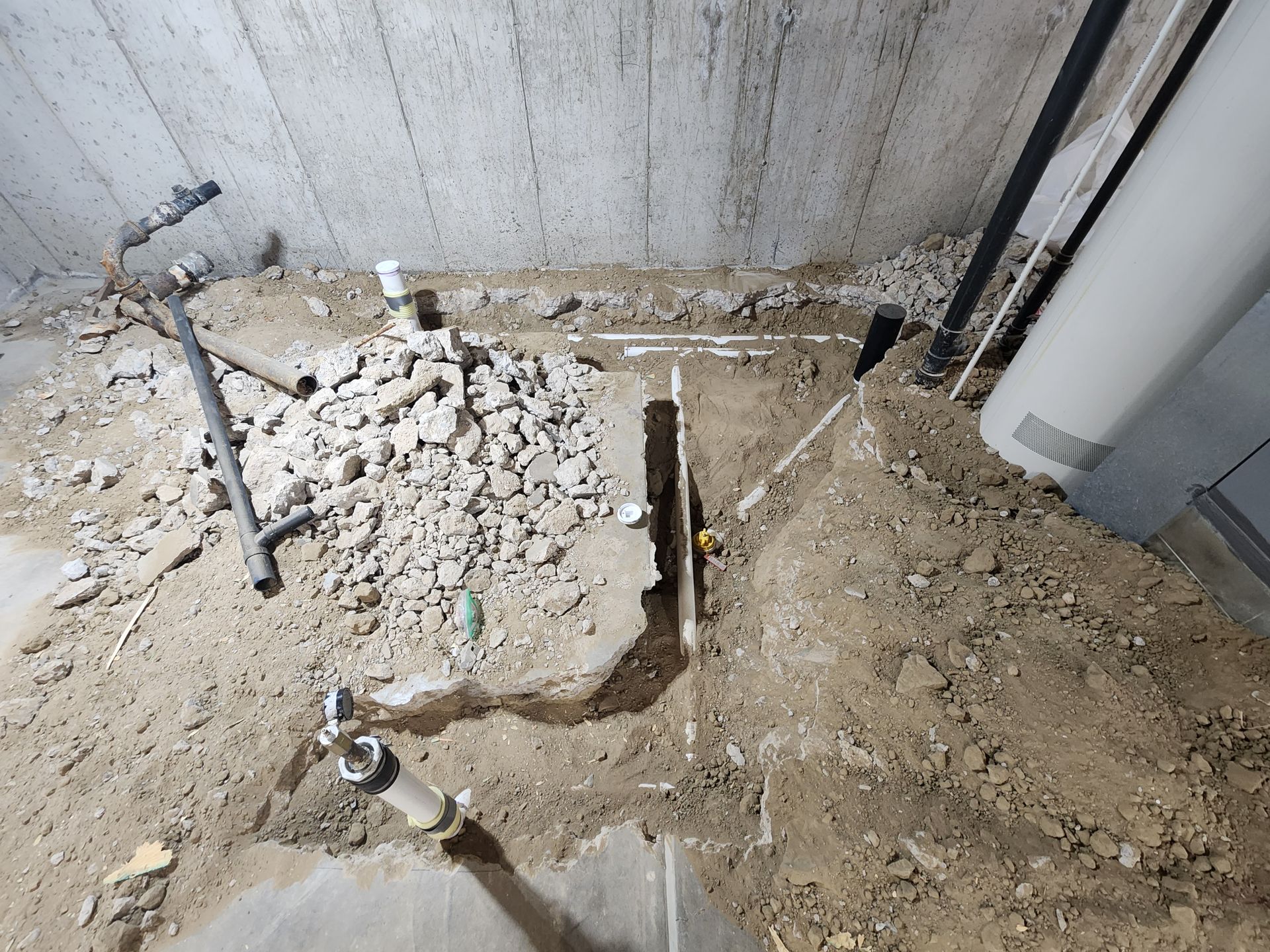 Construction site, exposed pipes in trench, rubble pile, dirt floor, concrete wall.
