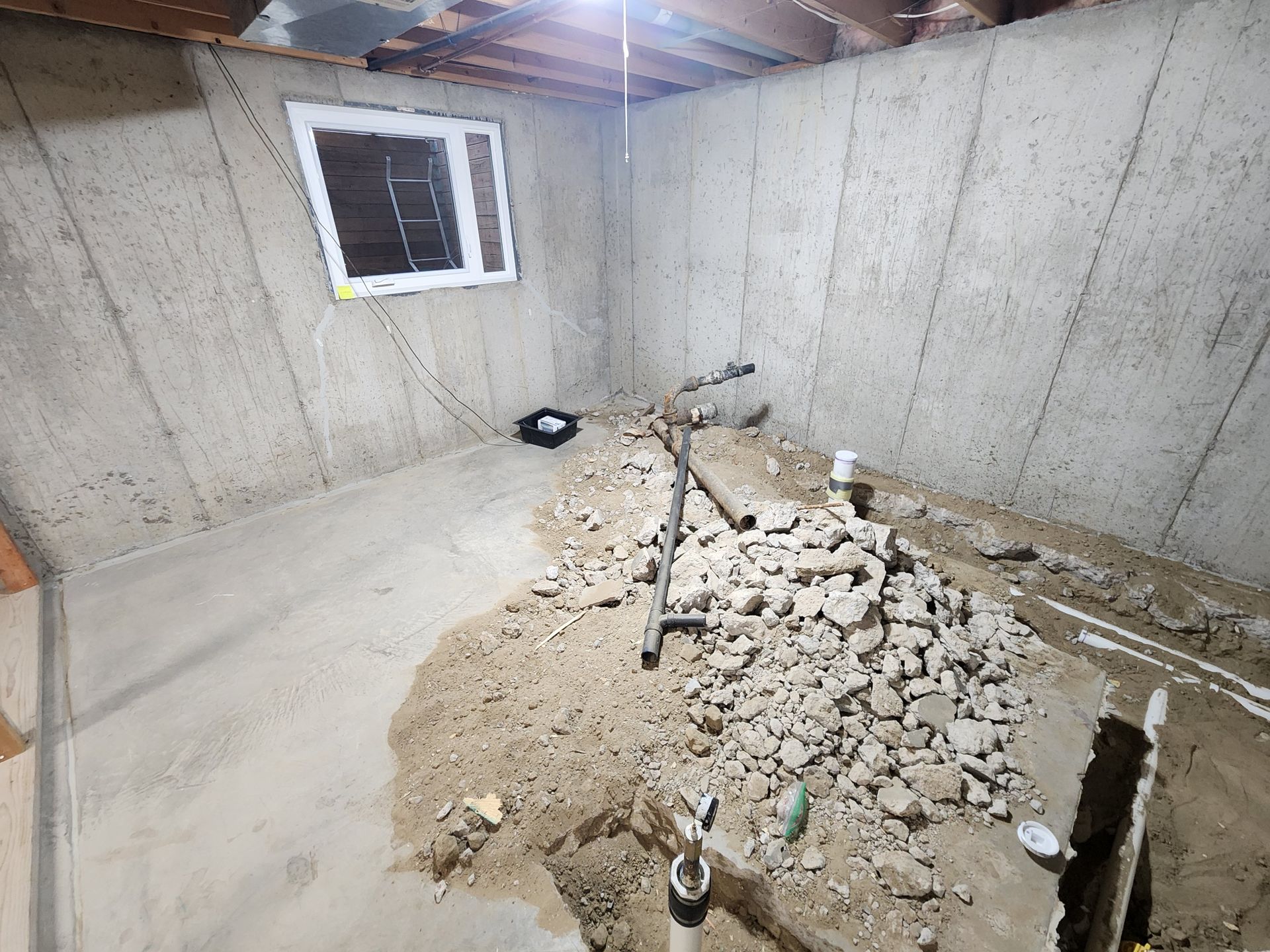 Basement renovation in progress: concrete walls, rubble on the floor, window, exposed pipes.