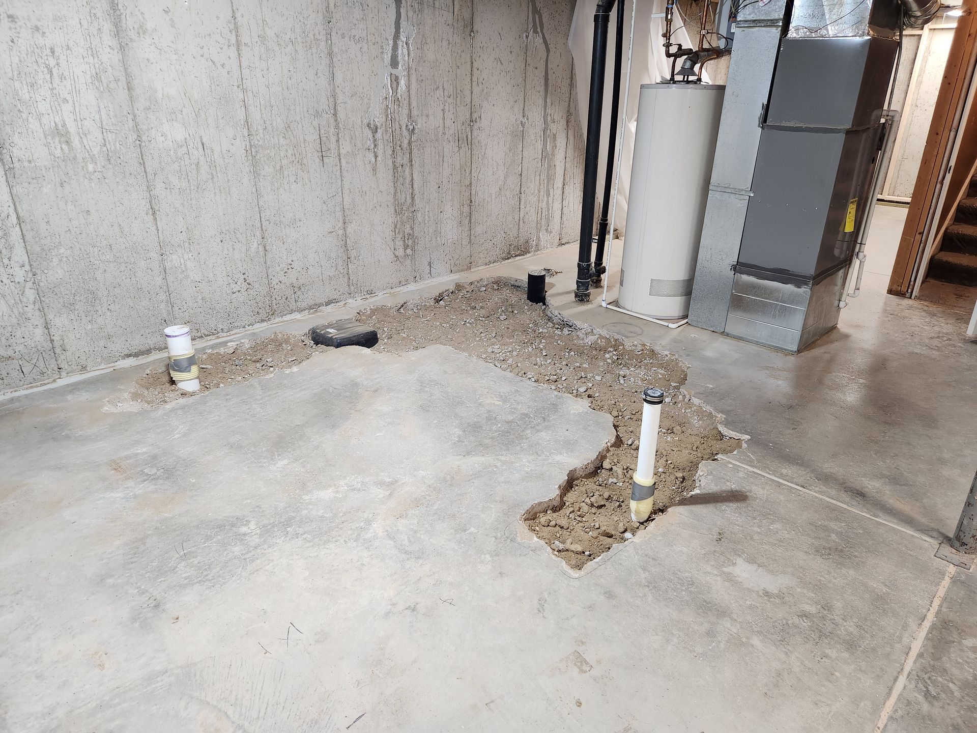 Basement with exposed pipes and gravel in concrete floor, near water heater, furnace, and stairs.