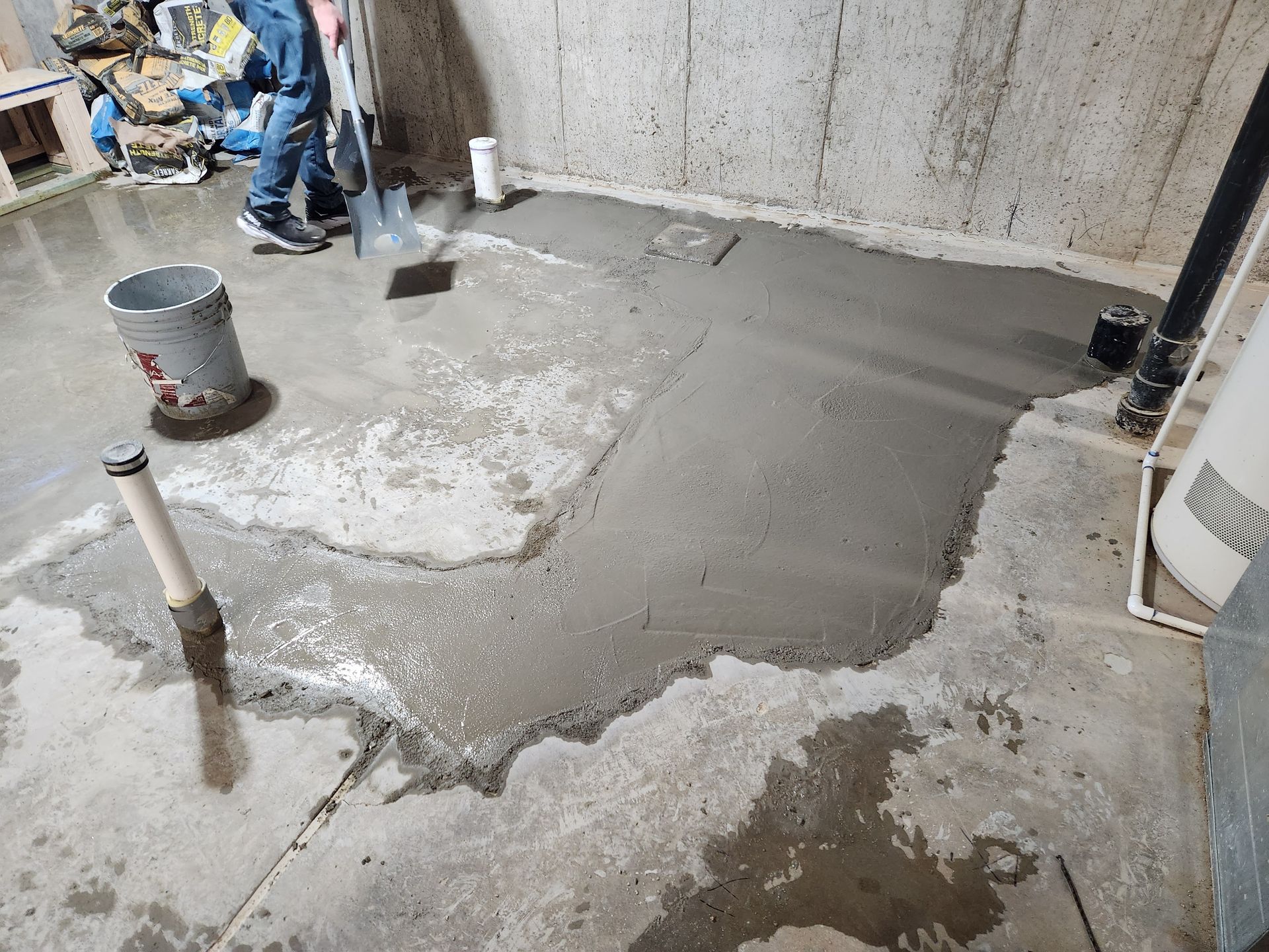 Person smoothing wet cement on a basement floor; includes buckets, pipes, and tools.