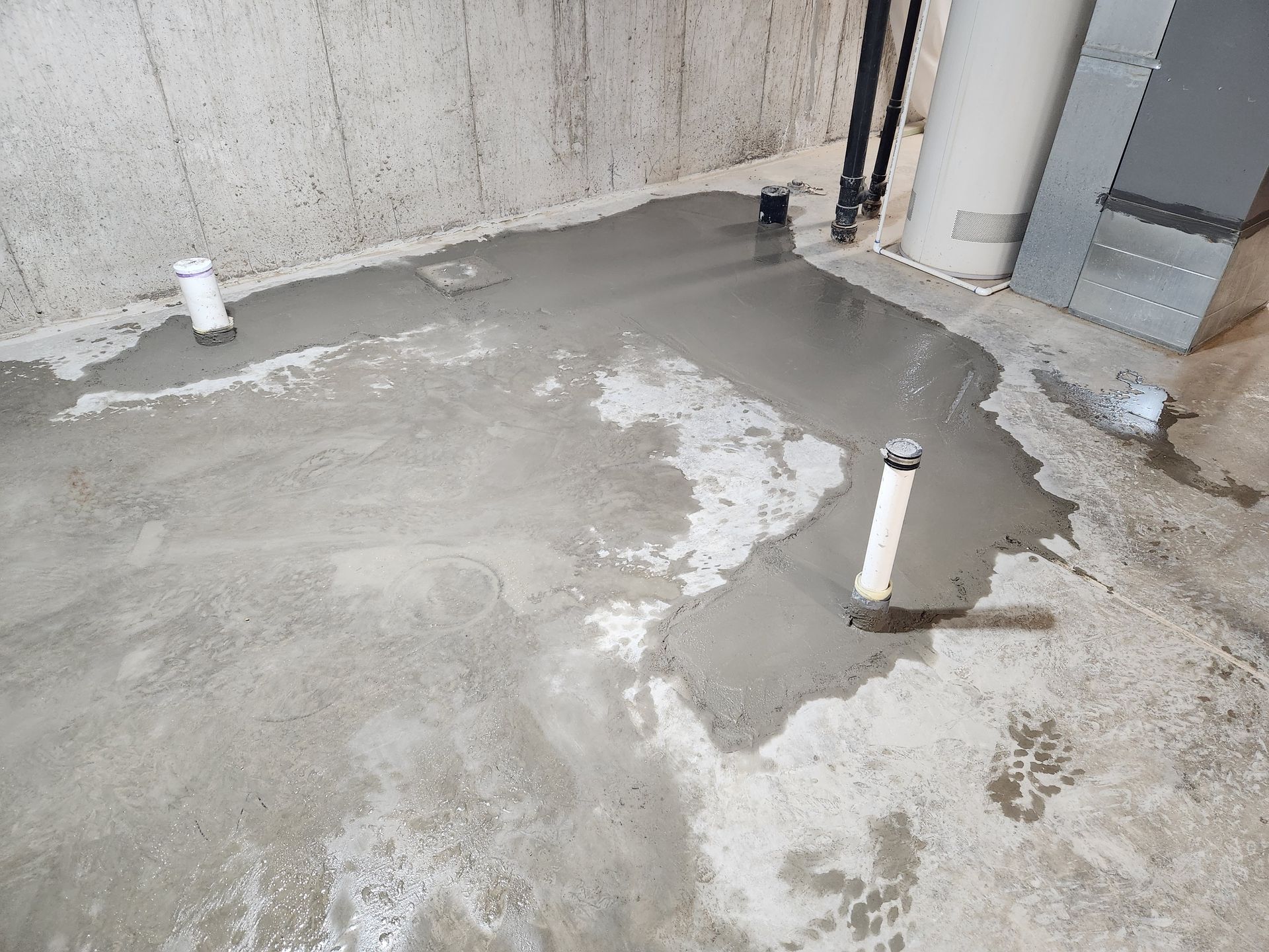 Basement floor with fresh concrete around pipes. Gray concrete and pipes against a light gray wall.