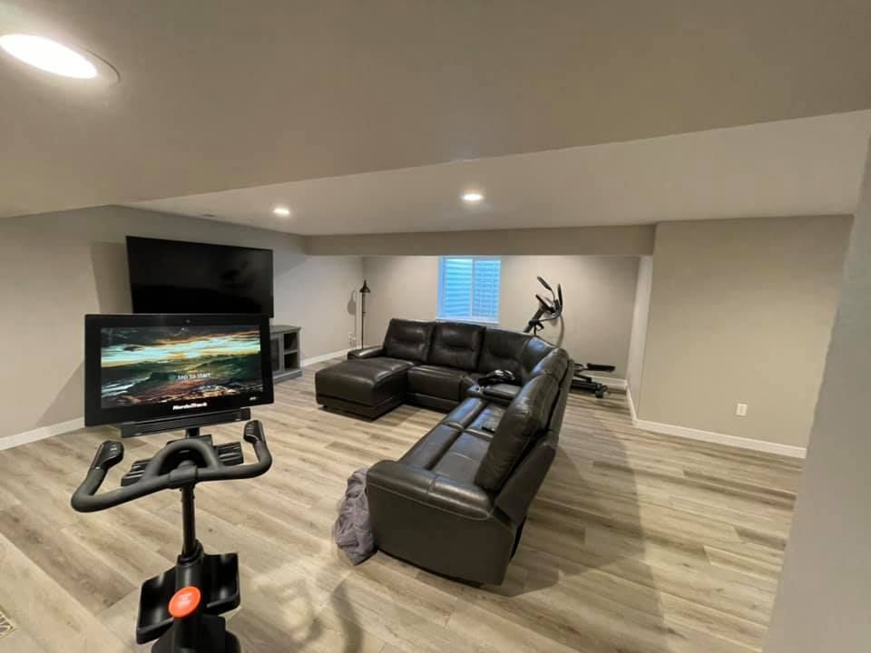 Basement living area with exercise bike, black sectional sofa, and TV on wall.