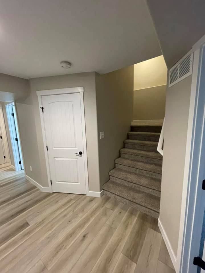 Basement with stairs, door, and light gray walls. Gray carpet on stairs, light wood flooring.
