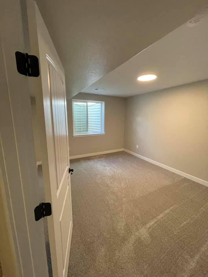A room with beige walls, carpet, small window, and a white door with black hinges.