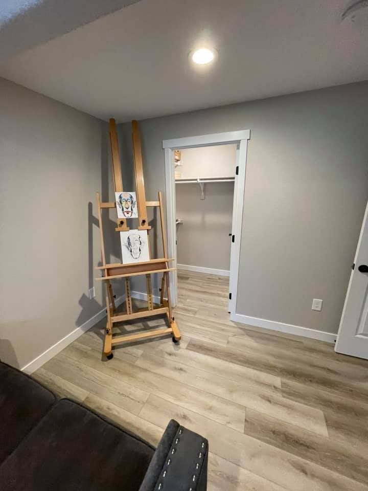 An art studio with an easel, artwork, and a doorway leading to a closet. The room has gray walls and wood flooring.